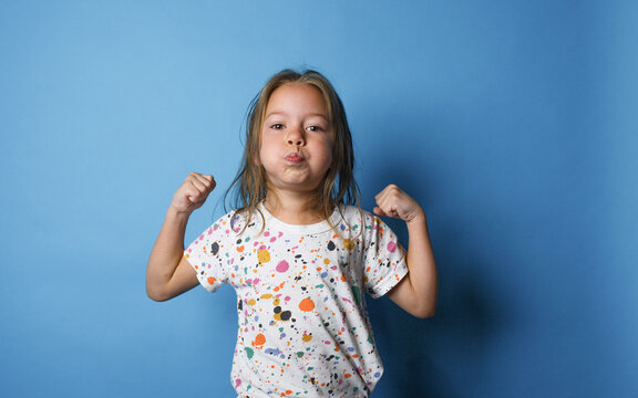 A Little Girl Shows Strength By Raising Her Arms Up And Puffing Out Her Cheeks On An Isolated Blue Background.