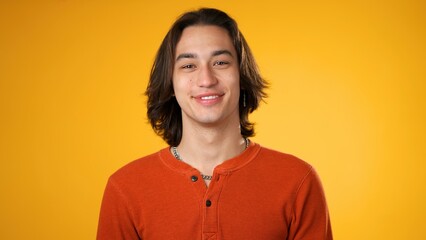 Smiling handsome Hispanic young man 20s wearing orange casual shirt isolated on yellow color background in studio. Portrait of gender fluid male sincere emotions lifestyle concept. Looking camera