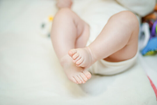 Crossed legs of the baby on a white background. A little girl lies on a white bed in a white bodysuit.