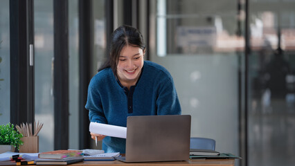Young Asian businesswoman standing at her office desk and working on her laptop with happiness.
