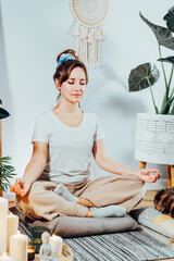 Young healthy woman practicing meditation at home, sitting in lotus position on floor cushion in...