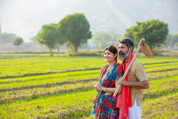 Indian farmer holding spade in hand and standing with wife at agriculture field.