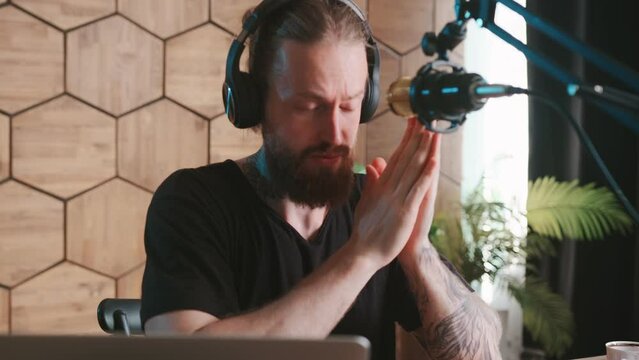 Young sensible restrained Caucasian man blogger recording audio podcast with serious face using professional studio microphone and headphones sits at desk in home office. Radio host, influencer