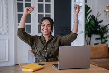 Surprised young pretty woman sitting at table rising hands, throwing paper sheets in air, happy to...
