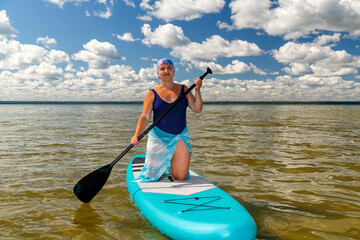 A Jewish woman in a kerchief kisuy rosh in a pareo on her knees on a SUP board swims in the lake on a sunny day.