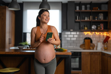 Pregnant woman in a maternity cloths with a beautiful big smile holding a smart phone in the...