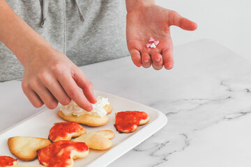 Hands of a caucasian teenage girl laying coffee cups heart sprinkles on not perfect freshly baked heart cookies .