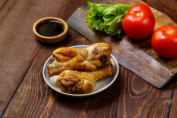 Baked chicken drumsticks on a plate on a wooden table next to soy sauce, herbs and tomatoes.