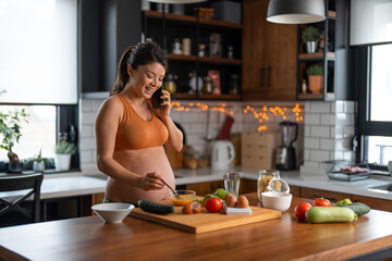 Obraz na płótnie Canvas Pregnant brunette mixing eggs and talking on the smartphone while standing at the kitchen counter.