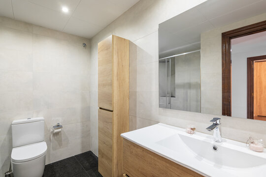 Modern Bathroom With White Tiled Walls. Small Wooden Cabinet For Toiletries And Towels Sits Against Wall And Harmonizes In Color With Cabinet For Sink. Mirror Reflects Doorway Into Room.