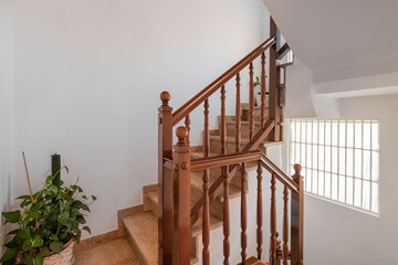 Spacious staircase with marble steps and wooden railings leading to upper floor against backdrop of white walls. Bright daylight from window well illuminates stairs and potted plant on floor.