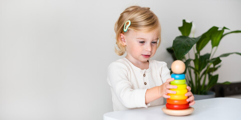 Adorable blonde toddler, kid, child, girl two years old playing indoor with eco wooden coloured toy pyramidion on white, gray background. Copy space.Early preschool development, kindergarten game.