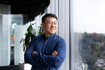 Obraz na płótnie Canvas Portrait of successful smiling businessman, Asian man with wrinkled hands in casual shirt looking out window, mature man in glasses working inside modern office.