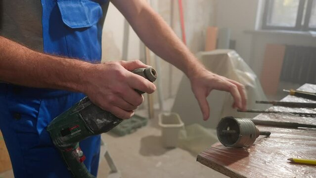Man in blue construction overalls puts nozzle on perforator to drill hole for socket. Builder installing round nozzle and checking its operation. Close up.