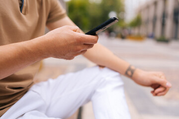 Close-up cropped shot of unrecognizable tattooed man sitting on bench using mobile phone looking to screen on city street. Unknow male in fashion clothes reading message on phone on summer day.