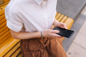 Close-up high-angle view of of unrecognizable young woman holding smartphone and typing online message with both hands sitting on yellow bench on city street. Unknown female holding cellphone in hands