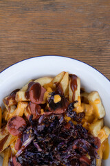 Top view of fried potatoes with sausages, red cabbage, cheddar cheese and barbecue sauce, in a white bowl with a wooden background.