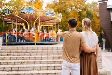Rear view of unrecognizable loving young couple standing embracing watching rotation of carousel in amusement park. Happy man pointing hand and woman walking spending time together on warm summer day.