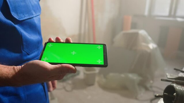 Male builder holding touch screen smartphone with green screen chroma key. Man using finger for touching, clicking, swiping, sliding, zooming, dragging on phone screen. Close up.