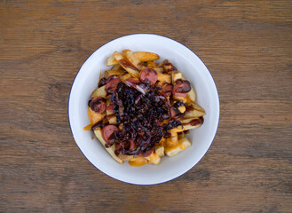 Top view of fried potatoes with sausages, red cabbage, cheddar cheese and barbecue sauce, in a white bowl with a wooden background.