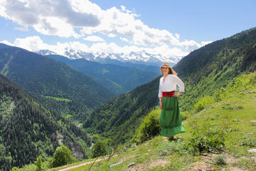 Fototapeta na wymiar Elegant woman in green skirt at the mountains. Barefoot on the grass. Happy girl with wavy hair with a hat. The background of the majestic mountains of the Caucasus and wooden fence. Svaneti, Georgia.