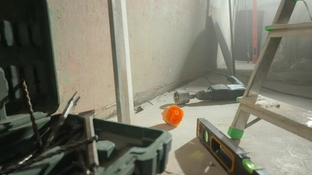 Perforator with nozzle for cutting holes for socket lying on floor next to toolbox and drills. Interior of the room inside is in process of renovation with ladder, boxes and level. Close up.