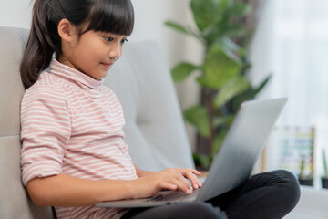 Asian Little school kid girl use laptop computer sitting on sofa alone at home. Child learning reading online social media content, play education lessons game chatting with friends. 