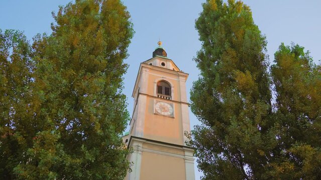 View on church historical bell tower building with clock on wall standing between high trees under clear sky in Ljubljana