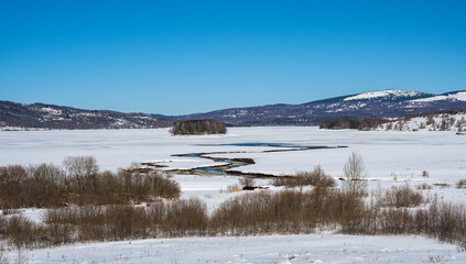 Winter landscape with a watch post and tributary River entering the frozen lake with with lake island and hills in the background background. Frozen Lake on a cold winter day. © Bojan