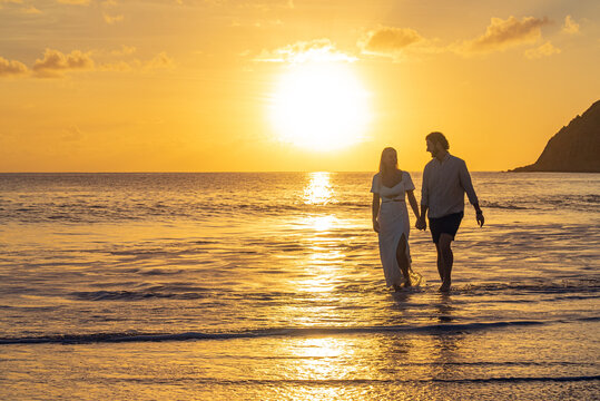 Dressed up young romantic couple walking on sandy tropical beach during the sunset over the ocean