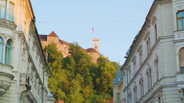 Historical castle with towers on hill top surrounded by large city park behind old buildings with monograms and balconies on Ljubljana street