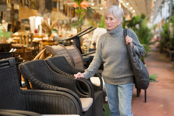 Positive aged female shopper choosing resin wicker chairs for using in outdoor patio or garden in furnishing salon ..