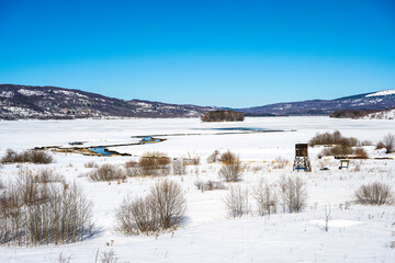 Winter landscape with a watch post and tributary River entering the frozen lake with with lake island and hills in the background background. Frozen Lake on a cold winter day.