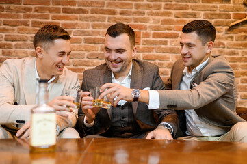 Front view of bridegroom and guys, wearing in suits, sitting on comfortable couch, holding glass with alcohol beverage and celebrating wedding in apartment