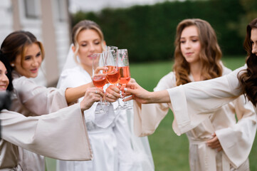 Front view of attractive bride and bridesmaids, standing together in circle, clinking glasses with pink wine and toasting while having party in wed morning