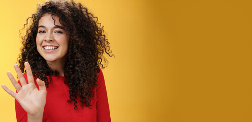 Charming friendly and self-assured attractive curly woman waving cute with palm to say hi or hello smiling broadly greeting man trying flirt in party posing joyful over yellow background