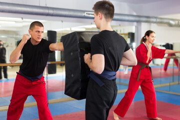 Fototapeta na wymiar Focused sporty man learning to punch in martial arts training, exercising on kick shield held by young assistant in gym