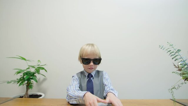 Blonde caucasian boy in smart suit and tie and too big sunglasses is looking into the camera, someone is giving him a bunch of banknotes. Someone pays little worker for his job. Businessman