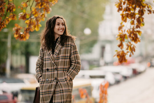 Side view of happy girl with dark hair, dressed in plaid coat and holding hands in pocket while walking on city street during autumn weather and looking away