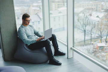 A young manager man in glasses and casual clothes sits on a soft chair and works on a laptop. Work...