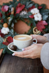 Woman hands holding cup of coffee in front of stylish Christmas wreath outside. Cozy festive home. New Year morning