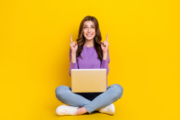 Full body photo of cheerful lady sit floor use netbook look indicate fingers up empty space isolated on yellow color background