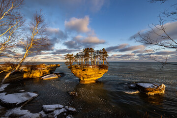 Sunrise illuminates this sculpted formation known as Turnip Rock on the Lake Huron shoreline in early winter. Pancake ice can be seen floating in the surrounding waters. Blue sky, puffy clouds.