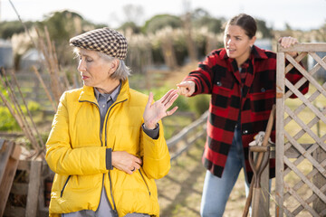 Two upset young and old female neighbors disagree with each other while standing near fence on...