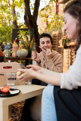 Happy young woman customer enjoying tasting organic fruits and veggies at local food market on sunny autumn day. Female farmer sitting at table with consumer, offering to taste homegrown produce