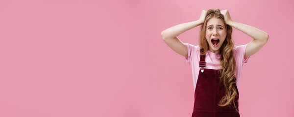 Waist-up shot of woman in panic screaming and pulling hair out of head grimacing feeling anxiety from stress and problems standing bothered and distressed against pink background