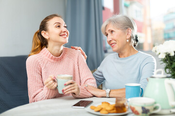 Two positive women, grandmother and her adult granddaughter, sitting at table and having conversation while drinking tea.