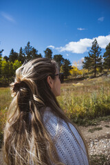 Woman looking out over a field pensively with golden aspens and pine trees - she has long blond hair and is wearing glasses and a white sweater