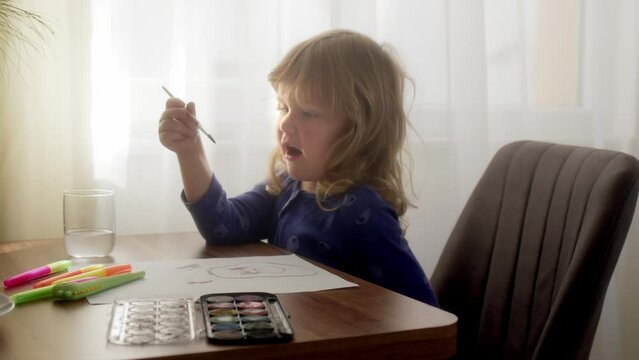 happy child paints with watercolors on paper at home or in garden. beautiful blonde girl with curly hair is having fun at table with paints and brush. Hobby kid child at home.