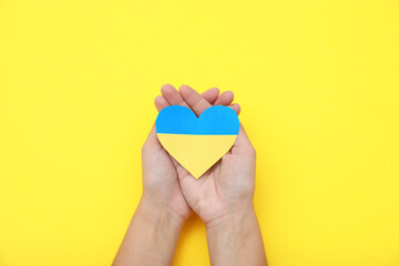 Fototapeta na wymiar Woman holding paper heart in colors of Ukrainian flag on yellow background, top view